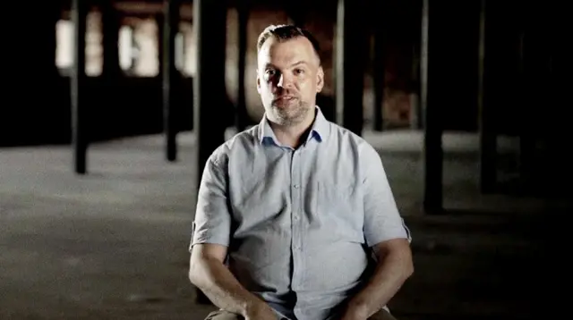 Иван Семенихин — мужчина средних лет с короткими светлыми волосами и лёгкой щетиной сидит в полутёмном помещении с кирпичными колоннами. На нём светло-голубая рубашка с короткими рукавами, он улыбается.
