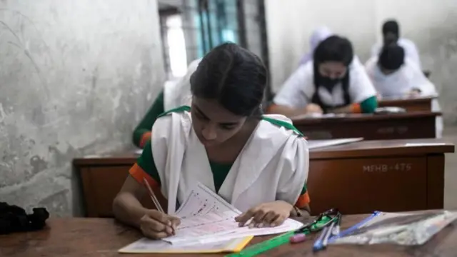 সবুজ ও সাদা স্কুল ড্রেস পরা এক ছাত্রী পরীক্ষার খাতায় বৃত্ত ভরাট করছে কলম দিয়ে, পেছনে আরো কয়েকজন পরীক্ষার্থী লিখছে