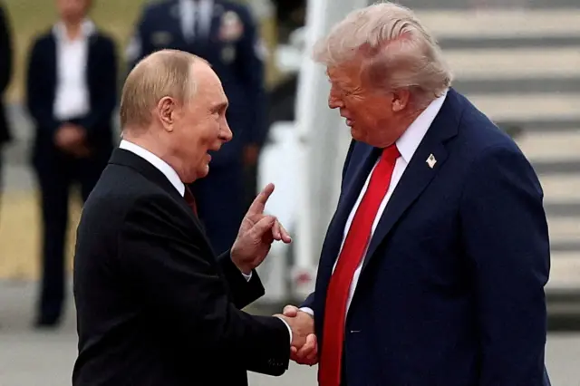Putin y Trump se dan la mano frente a la escalerilla de un avión. Putin levanta la mano izquierda con el dedo índice apuntando hacia arriba y habla con Trump con la boca abierta. Hay gente al fondo; uno de ellos lleva condecoraciones militares. Trump viste una chaqueta oscura y corbata roja. Putin lleva una chaqueta negra. Ambos visten camisa blanca.