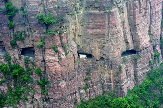 Pared de roca de una montaña con agujeros por los que se ve un camión transitando la carretera esculpida