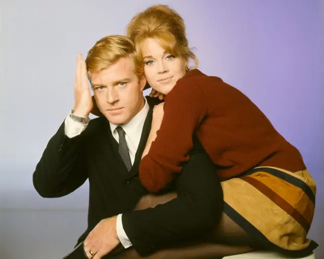 Jane Fonda vestido con traje formal oscuro y corbata abrazado por Jane Fonda quien está vestida con un saco rojo oscuro y una falda amarilla con rayas negras y rojas