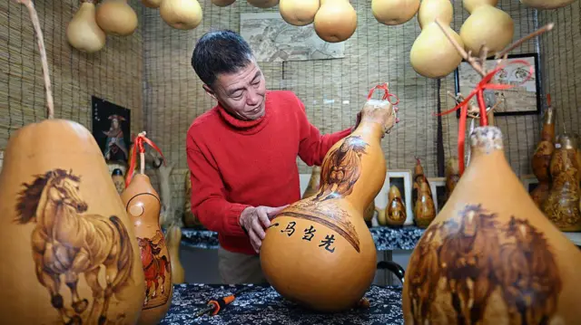 Um homem vestido com um suéter vermelho observa uma série de objetos com imagens de cavalos pintadas neles.