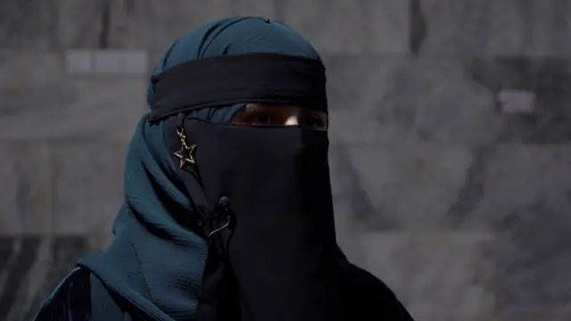 Safia, le visage couvert, dans une salle de classe, devant un mur.