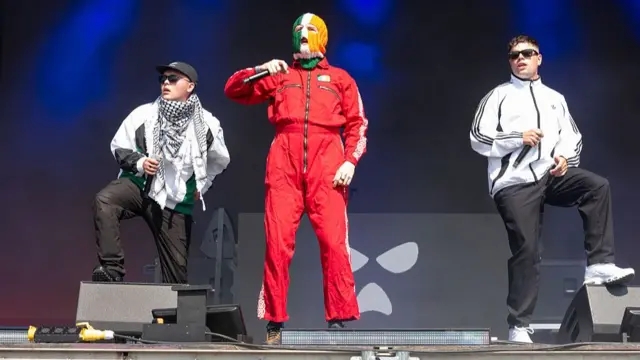 ニーキャップの3人のメンバーがステージに立っている。中央の人物は、赤いライダースーツを着て、アイルランドの国旗のオレンジ・白・緑に色分けされたバラクラバを被っている。右側の人物は白い上着に黒いパンツ姿で、サングラスをかけている。左側の人物は白い上着、黒いパンツ、サングラス、黒のキャップという姿で、首にパレスチナの伝統的な布地「クフィーヤ」を巻いている