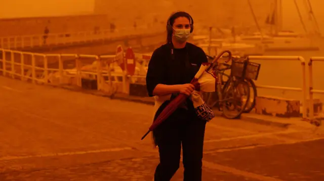 Девојка носи кишобран, има маску преко лица и велике слушалице