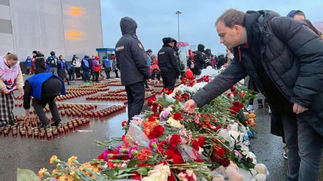 Pessoas colocando flores no chão e acendendo velascódigo promocional betano benficahomenagem às vítimas