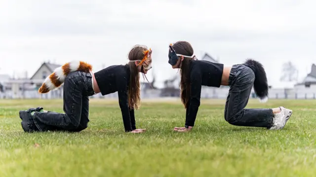Dve tinejdžerke sa maskama životinja na licu i repovima kleče oslonjene rukama na zemlju