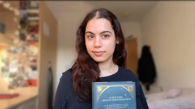 Ciemnowłosa kobieta w czarnej bluzce. Patrzy w kamerę, w ręku ma książkę o niebieskiej okładce. W tle widać biurko, szafę i łóżko.