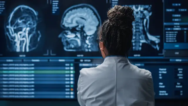 A neurologist stands with her back to the camera, looking at brain scans on a large screen in front of her.