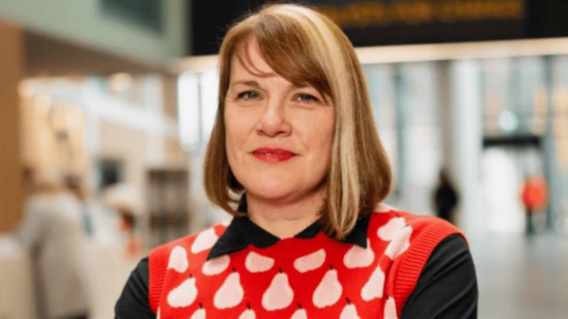 Primer plano profesional de la Dra. Jane Meyrick con una camisa negra de cuello bajo un chaleco rojo de punto con peras blancas. Su cabello es castaño rojizo con un mechón rubio, cortado a la altura de los hombros y con flequillo lateral.