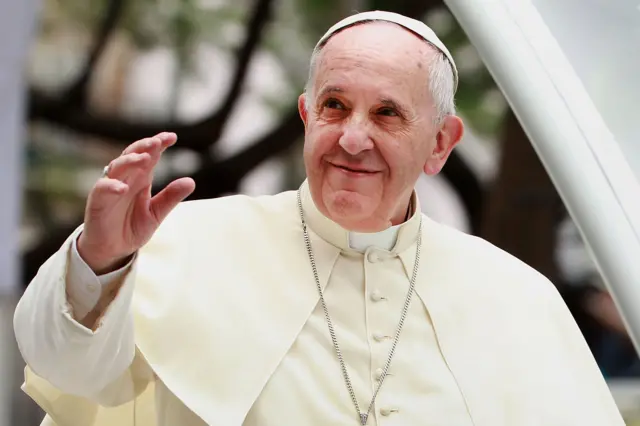 Pope Francis is wearing a cream-coloured robe with a matching skullcap and a long silver chain with a cross pendant. One hand is raised in a gesture, while the other is partially visible. The background shows blurred greenery and architectural elements, suggesting an outdoor setting at a formal or ceremonial event.