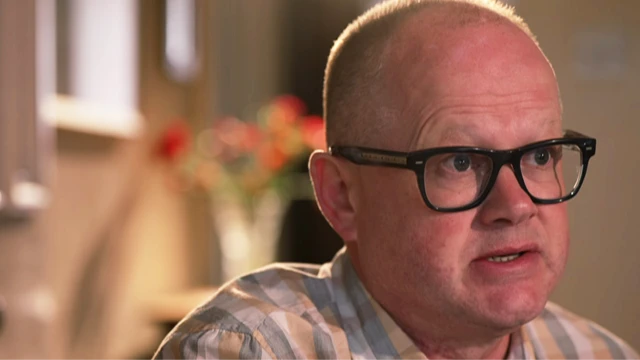 Marc Edwards is a white man with balding, fair closely cropped hair. His head and shoulders are visible. He wears black, heavy-rimmed glasses and a grey, white and tan checked shirt. He is looking towards the right of the image and appears to be talking seriously. The background is blurred but it is a room and a vase of flowers on a shelf can be discerned.