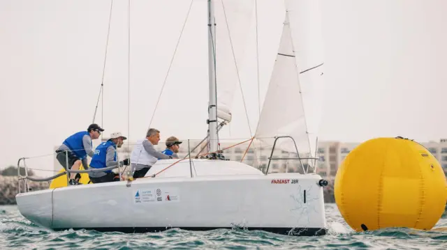 Karl Haines está a la izquierda de la imagen con una gorra negra, junto con los otros miembros de la tripulación del equipo GBR con discapacidad visual, compitiendo alrededor de una gran boya amarilla, en el Campeonato Mundial de Inclusión de Vela en Omán.