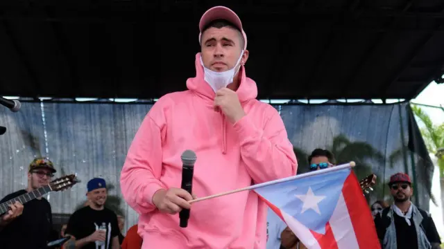 Bad Bunny con sudadera rosa y un micrófono sostiene una bandera de Puerto Rico con el triángulo celeste.