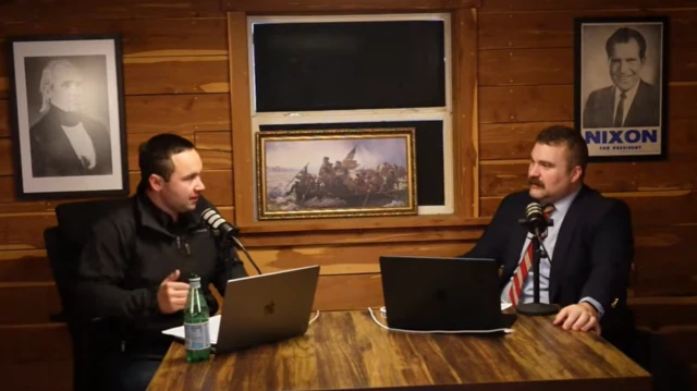 Dois homens sentados frente ao microfone e computadores, com imagens consideradas patrióticas ao fundo. Entre elas, inclui-se uma cópia de um famoso quadro de George Washington durante a Guerra da Independência dos Estados Unidos e fotos dos ex-presidentes Richard Nixon e James Polk.