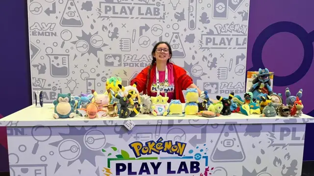 Kırmızı ceketli, esmer bir kız, rengarenk Pokémon peluş oyuncaklarıyla dolu markalı bir panonun arkasında gülümseyerek duruyor.