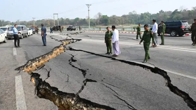 Several Myanmar military council staff look on at cracked roads in the capital after the massive earthquake. There are several cars in the background. 