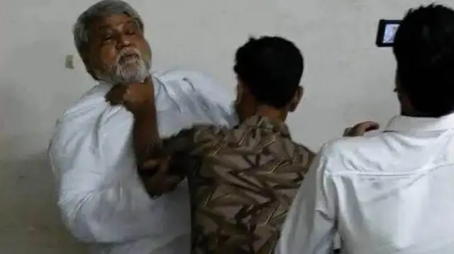 একজনের গলা চেপে ধরেছেন আরেক জন ব্যক্তি, পাশে তৃতীয় একজনের পিঠ দেখা যাচ্ছে