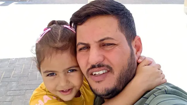 kahverengi saçları pembe tokalarla arkaya bağlı olan sarı tişörtlü Mira (solda), kollarını Saeed'in boynuna dolamış gülümsüyor. Saeed'in kahverengi saçları, kısa sakalı ve bıyığı var. İkisi de kameraya bakıyor.