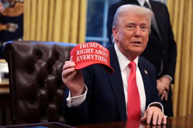 Trump no Salão Oval da Casa Branca segurando boné vermelho com a estampa "Trump estava certo sobre tudo" em inglês