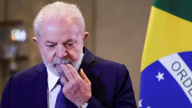 Foto mostra Lula com a mão esquerda levada à boca próximo a uma bandeira do Brasil 