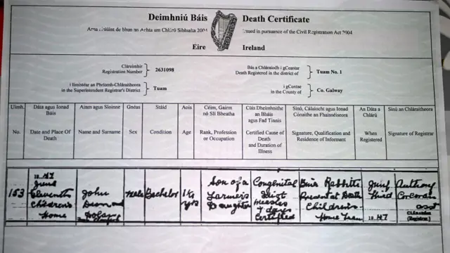 Primer plano de un certificado de defunción irlandés. Contiene una tabla de información que incluye nombre, fecha y lugar de fallecimiento, sexo, edad y otros detalles. La información está escrita en cursiva fluida y señala que el fallecido se llama John, tenía 16 meses y menciona "idiota congénito" y "sarampión" como causa de muerte.