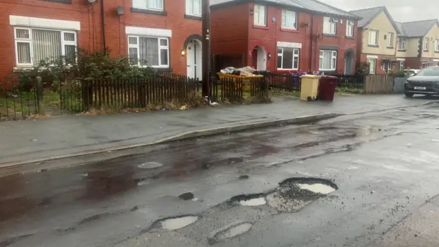 Fotografía de baches en la Avenida Ramsay, en la zona de Farnworth, Bolton. Al fondo se ven casas, contenedores de basura y coches.