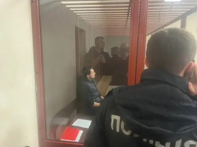 На задньому плані Володимир Кудрицький сидить боком до камери в скляному боксі суду. На передньому спиною до камери - поліцейський. 