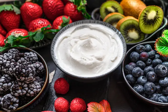 A bowl of yoghurt surrounded by bowls of blueberries, blackberries, strawberries and cut kiwi fruit,