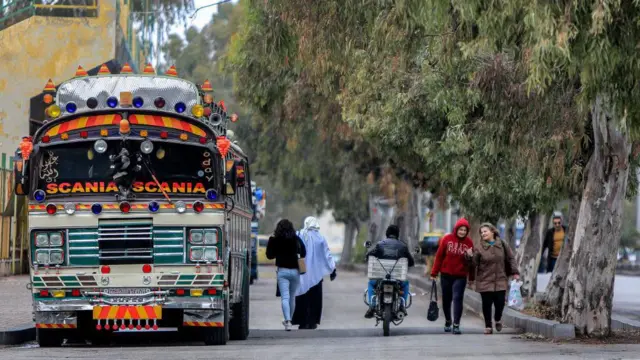 Mulheres caminham em rua movimentada de cidade síria, ao lado de ônibus decorado com lanternas e placas coloridas.