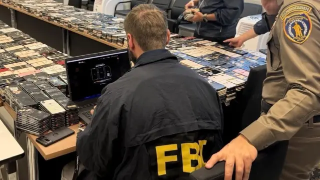 အဝါရောင် FBI တံဆိပ်ပါ ကုတ်အင်္ကျီဝတ်ထားတဲ့အမျိုးသားတစ်ဦး ကွန်ပျူတာရှေ့မှာ ကျောပေးထိုင်နေပြီး သူ့နောက်မှာ ထိုင်းရဲတပ်ဖွဲ့ဝင်တစ်ဦး ဘေးတစ်စောင်းနဲ့ မတ်တပ်ရပ်နေတာကို မြင်တွေ့ရ။ ရှေ့က ခုံတန်းရှည်ပေါ်မှာတော့ အဆင့်ဆင့်ထပ်ထားတဲ့ လက်ကိုင်ဖုန်းရာပေါင်းများစွာကို မြင်တွေ့ရ 