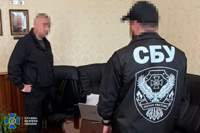 Приміщення зі столом і шкіряним чорним диваном уздовж стіни.
У кадрі двоє людей: чоловік із заблюреним обличчям стоїть біля столу, інший – повернутий спиною і в куртці з великим написом «СБУ» та емблемою на спині. На столі видно ноутбук із заблюреним логотипом.
