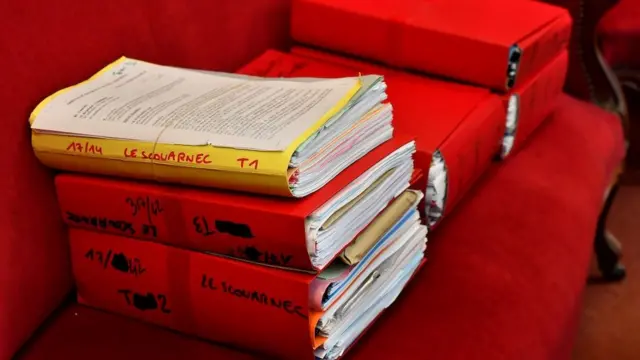 Red and yellow binders full of legal papers setting out the case against Joel Le Scouarnec in his 2020 trial. They are piled up on a plush red sofa.