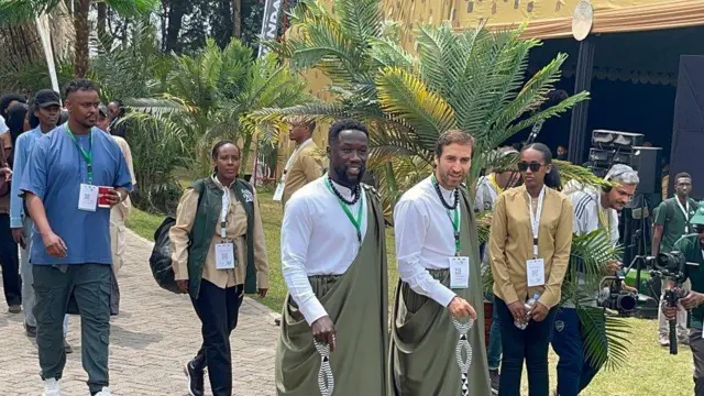 Abagabo babiri bambaye amashati y'umweru n'imikenyero ya Kinyarwanda y'icyatsi kibisi, bashinze hasi ingabo ziriho amabara y'umweru n'umukara. Barareba imbere. Bakikijwe n'abandi bantu, barimo n'abafite za 'cameras'.