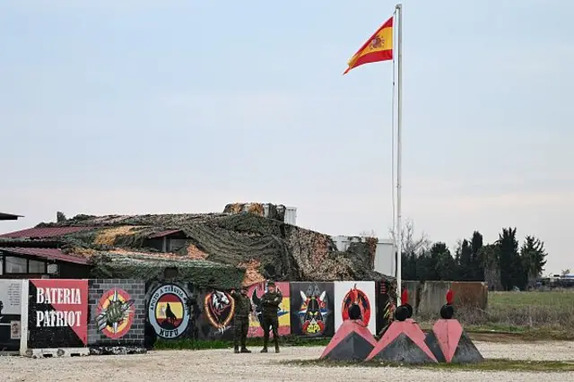 İspanya bayrağının dalgalandığı alanda iki asker konuluyor geride duvarda grafitiler var birinde Bateria Patriot yazıyor.