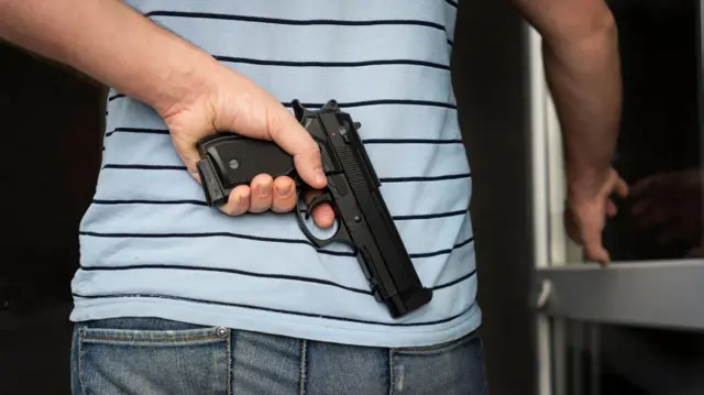 Un hombre esconde un arma en su espalda mientras abre una puerta