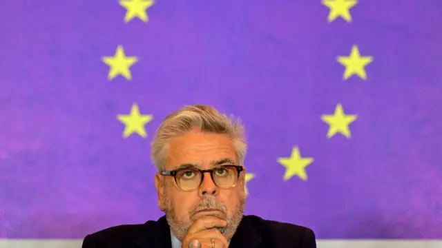 Sanchez Amor elini çenesinde tutuyor. Arkasında AB bayrağı var.