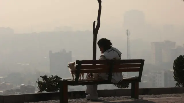 زنی نشسته بر روی یک نیمکت در تهران با هوای تیره و آلوده