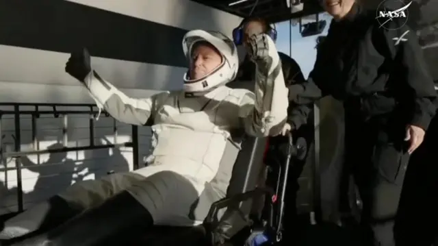 Butch erguendo os braços ao deixar cápsula, auxiliado por outras pessoas