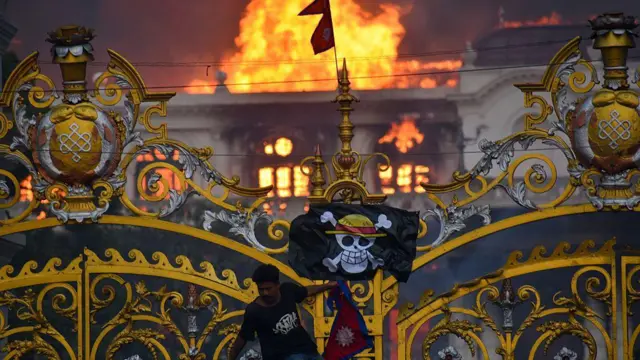 ساختمان پارلمان نپال در پس‌زمینه در آتش. یک معترض از دروازه ورودی آویزان است.