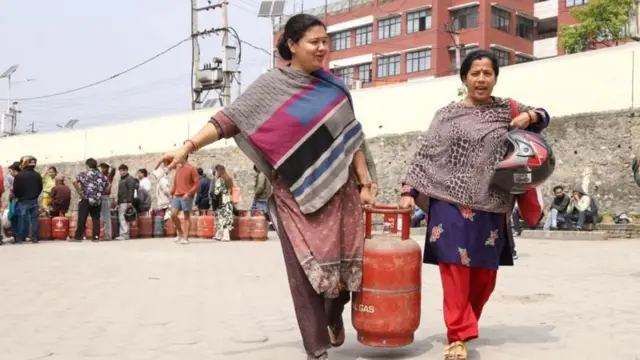 ग्यास सिलिन्डर बोकेर हिँडेका दुई महिला र पृष्ठभूमिमा ग्यास किन्न लाममा बसेका उपभोक्ताहरू