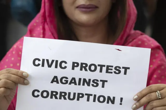 काठमाण्डूमा भ्रष्टाचार विरूद्धको प्रदर्शनमा एउटा कागजमा भ्रष्टाचार विरोधी नारा बोकेर सहभागी भएकी गुलाबी रङ्गको सल लगाएकी एक महिला।
