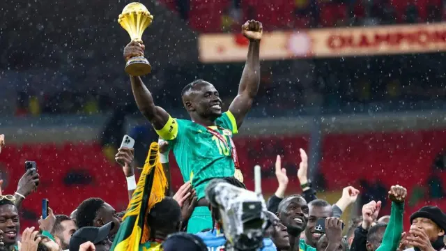 Le joueur sénégalais Sadio Mané, vêtu d’un maillot vert et portant une médaille dorée suspendue à un ruban rouge autour du cou, est porté par ses coéquipiers tandis qu’il lève les deux bras en l’air, tenant le trophée de la Coupe d’Afrique des nations dans sa main droite. Beaucoup de personnes dans la foule sous Mané le filment avec leurs téléphones portables et lèvent également les bras, tandis que des gouttes de pluie sont visibles sur fond flou de sièges rouges vides dans le stade de Rabat.