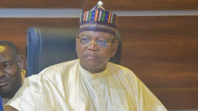 Sule Lamido sanye da babbar riga da yar ciki masu launin ruwon madara da hula mai launuka da dama da kula farin gilashi