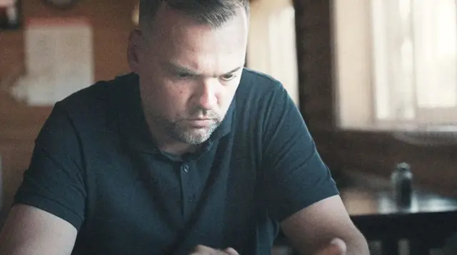 Иван Семенихин — мужчина средних лет с короткой стрижкой и сосредоточенным выражением лица сидит за деревянным столом в комнате с деревянными стенами и окном. На нем темно-синее поло, он смотрит в телефон. 