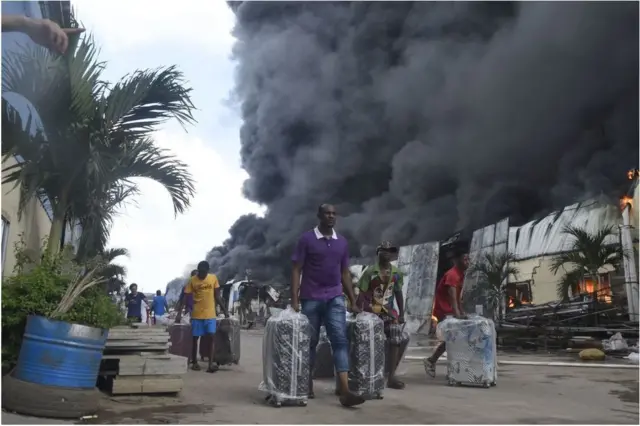 Abagabo barimo gukwega amavalisi mu muhanda inyuma yabo hari umwotsi mwinshi