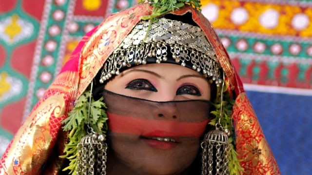 The image shows a bedouin woman dressed in elaborate attire, richly adorned with jewellry and textiles. The clothing is a vibrant red fabric with intricate gold patterns