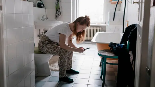 Mulher sentada no vaso sanitário, olhando o telefone celular