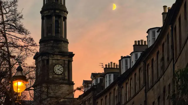 Una alta torre de reloj y una hilera curva de edificios al atardecer, con una luna creciente en el cielo y una farola brillante en primer plano.