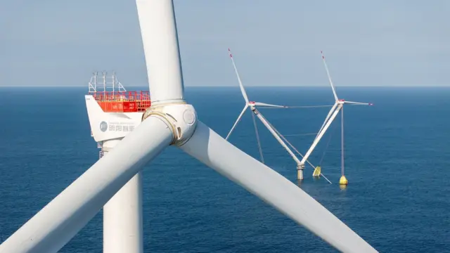 Plano cerrado del eje de las aspas de una turbina eólica marina blanca en el mar azul, con una torre de dos turbinas en el fondo 